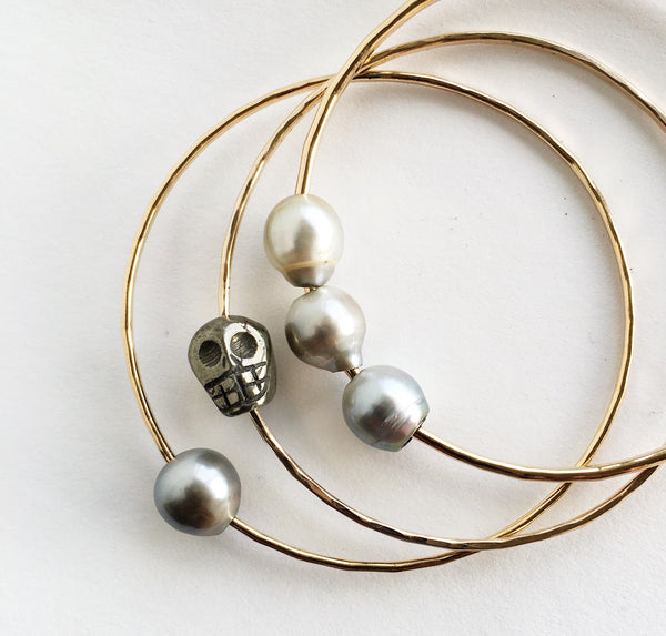 Carved pyrite skull bead slide across soldered bangle with hand-hammered texture. Displayed with single & triple Tahitian pearls handcrafted 12 gauge, 14KT gold-filled bangles.